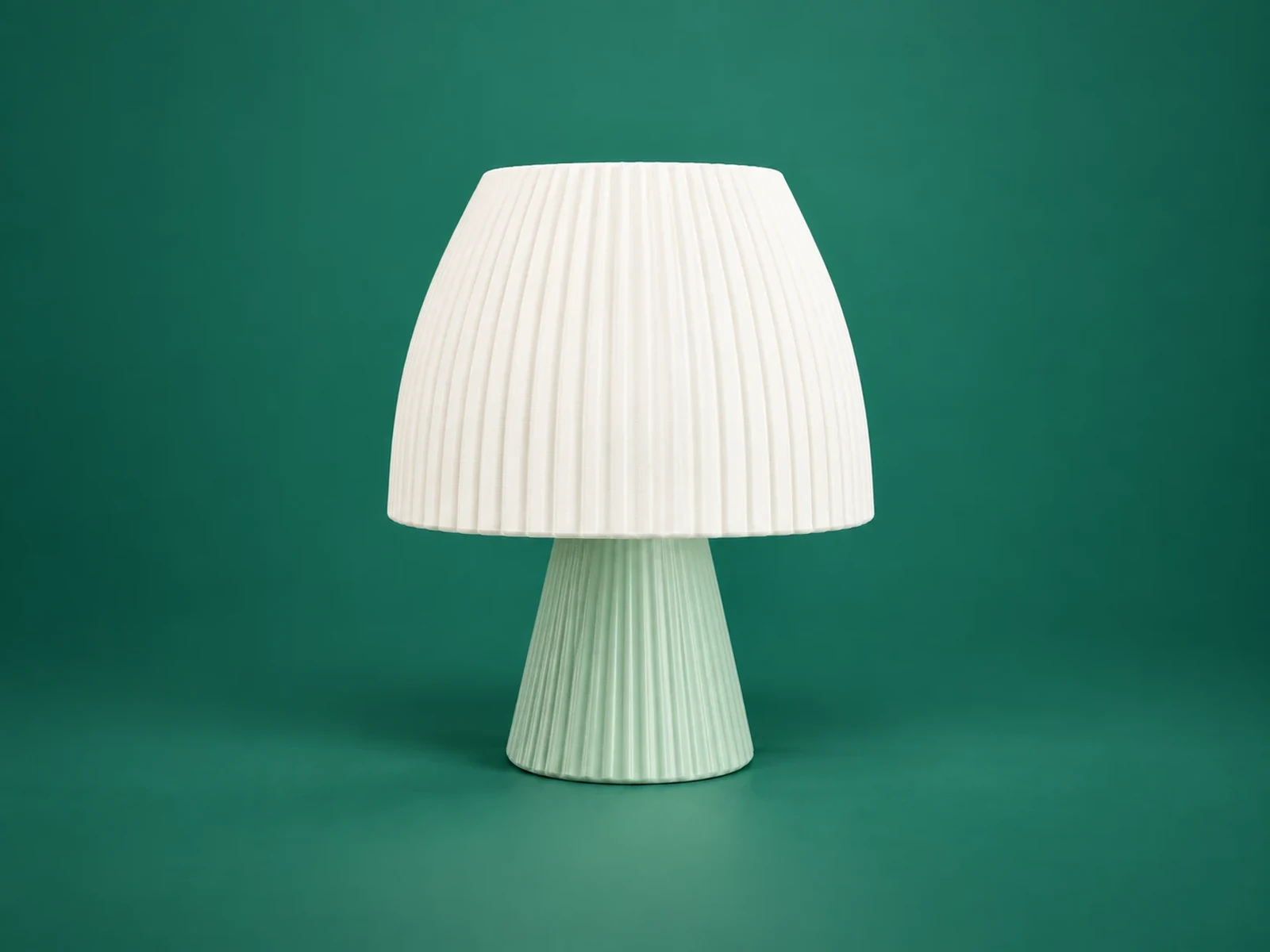 A 3D-printed table lamp with a white pleated shade on a mint fluted base, on a deep green background.