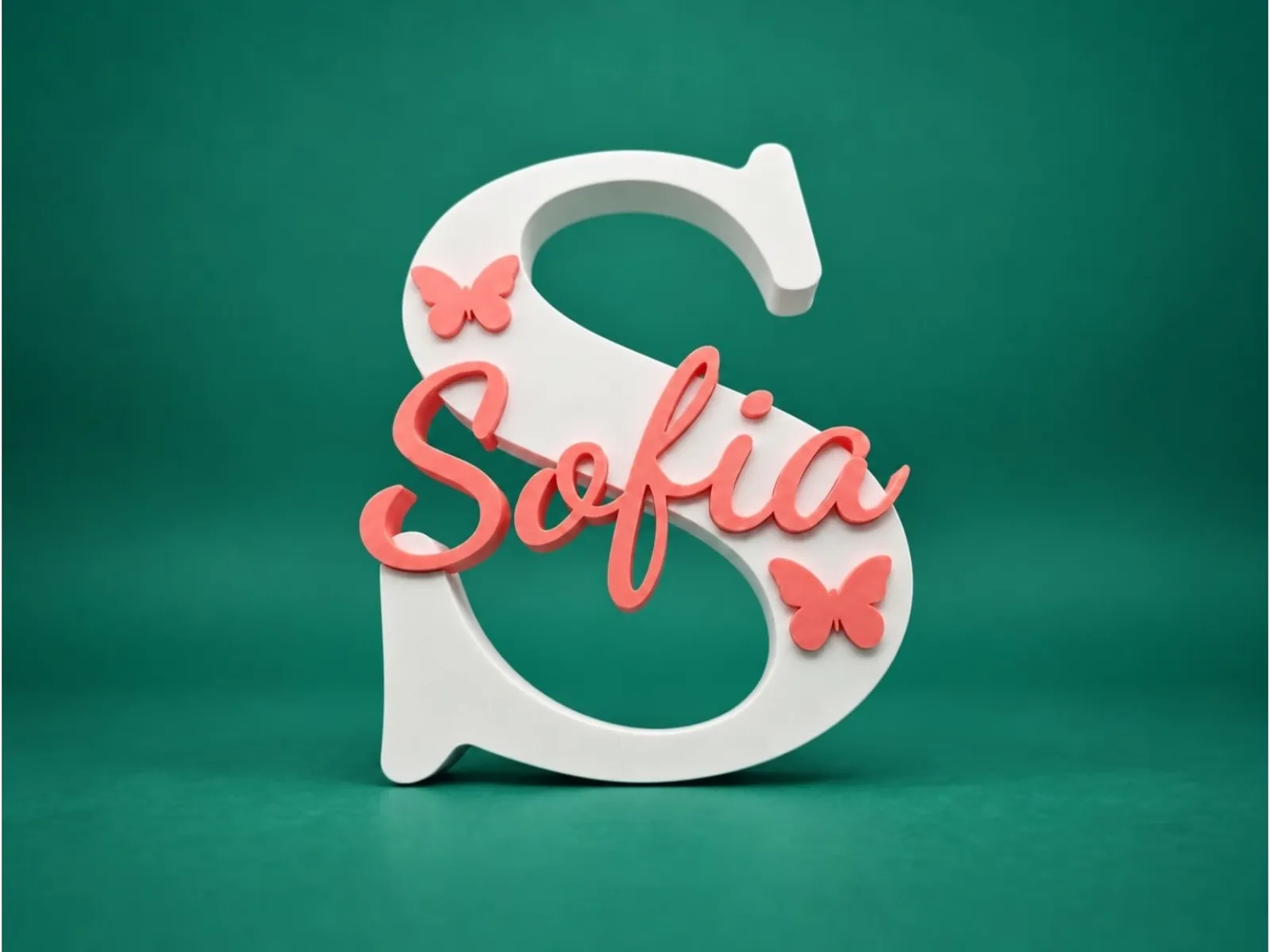 A white 3D-printed letter S with the name Sofia and two butterflies inlaid in coral pink, on a green background.