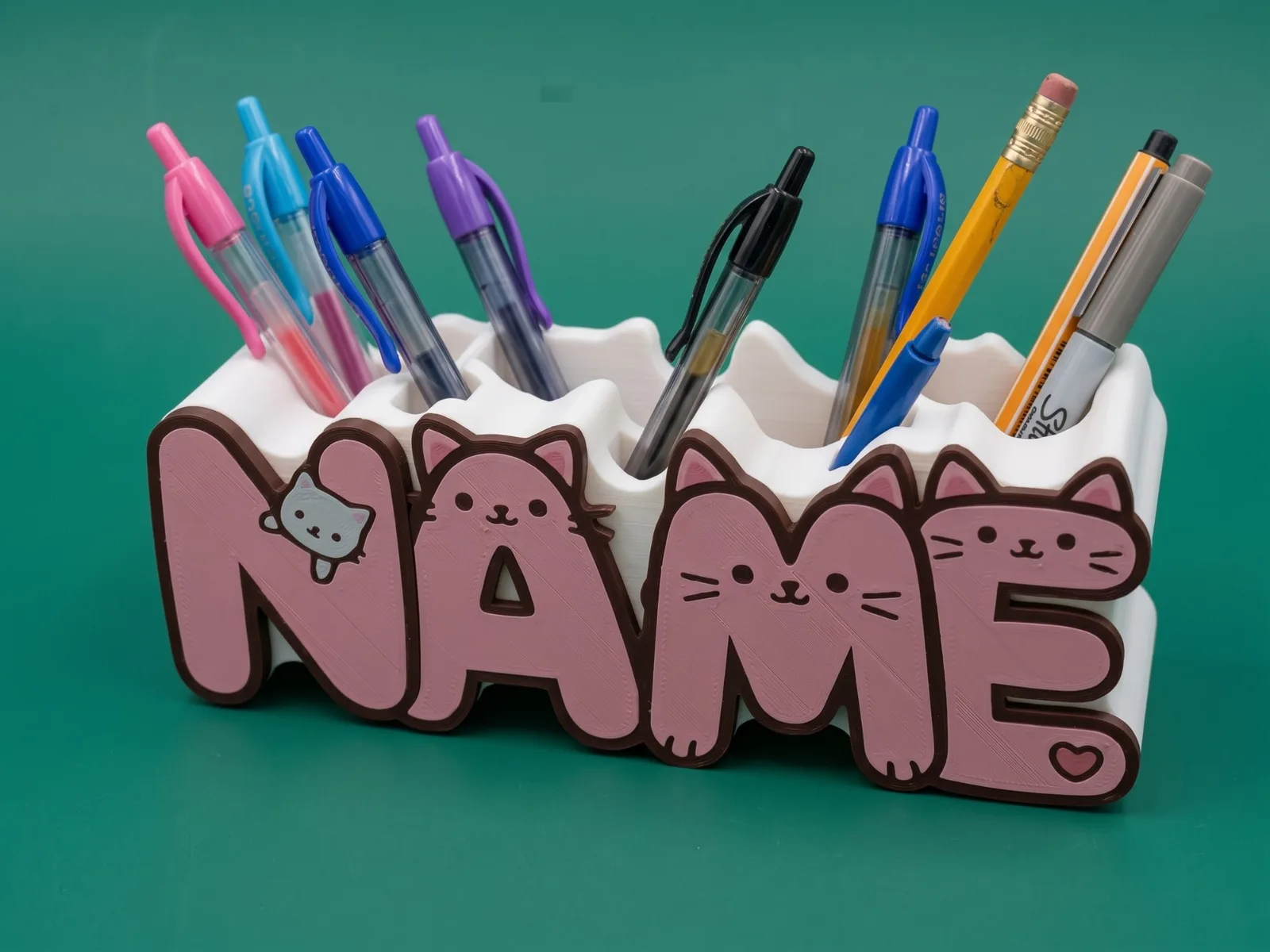 A 3D-printed name pen holder in the illustrated Kitty Pop font: the letters of NAME in pink with brown outlines and cat faces, in front of a white base holding colorful pens and pencils, on a green background.